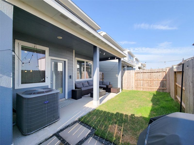 Cozy backyard patio area with a covered seating space, neatly bordered by a wooden fence. It features a small grassy area and is equipped with an outdoor air conditioning unit. Perfect for relaxing and entertaining.
