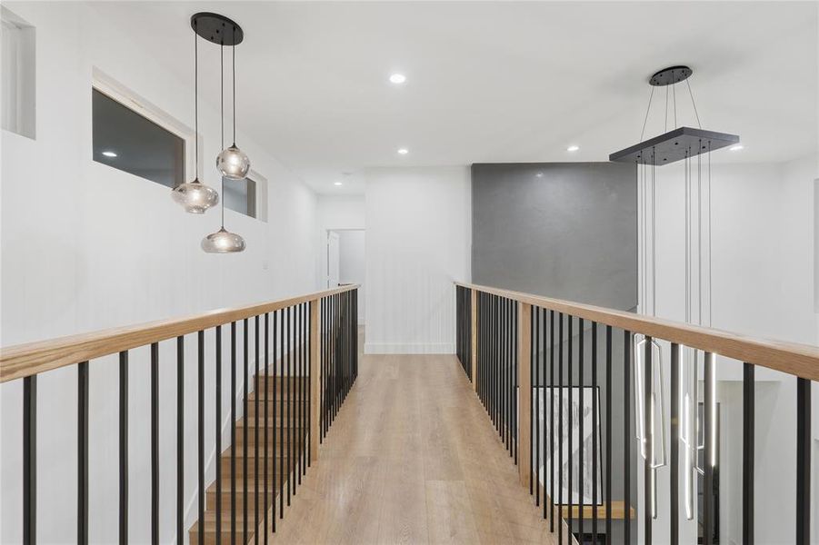 Upper-level hallway featuring wood-finish flooring, contemporary lighting fixtures, a light wood handrail with black iron balusters, recessed ceiling lights, and transom windows Upper-level hallway featuring wood-finish flooring, contemporary lighting fixtures, a light wood handrail with black iron balusters, recessed ceiling lights, and transom windows