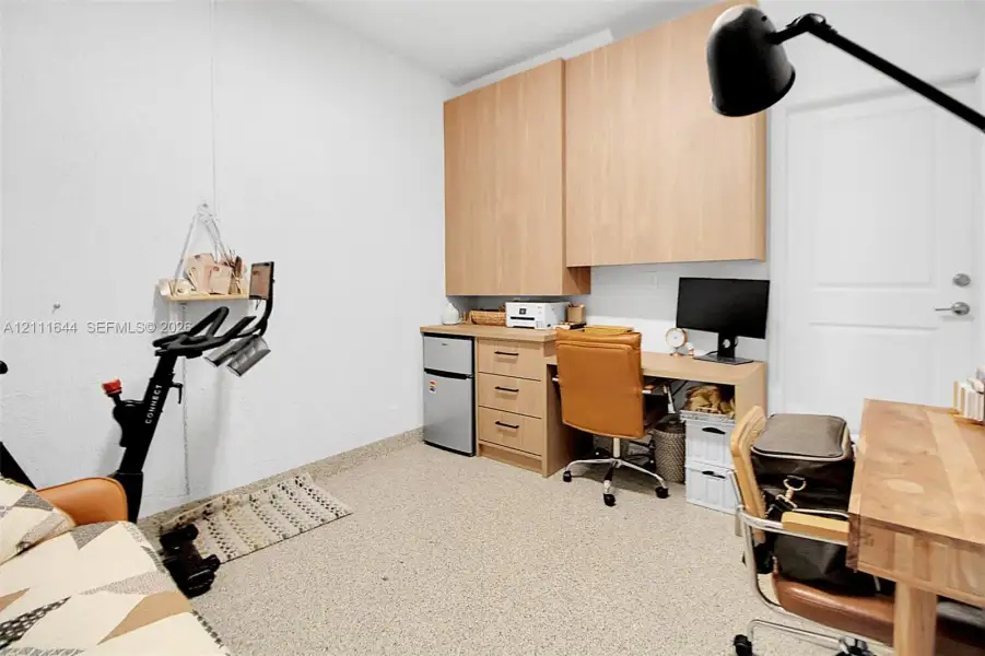 Built-in light-wood cabinetry with upper storage, integrated desk workspace with three drawers, and textured flooring Built-in light-wood cabinetry with upper storage, integrated desk workspace with three drawers, and textured flooring