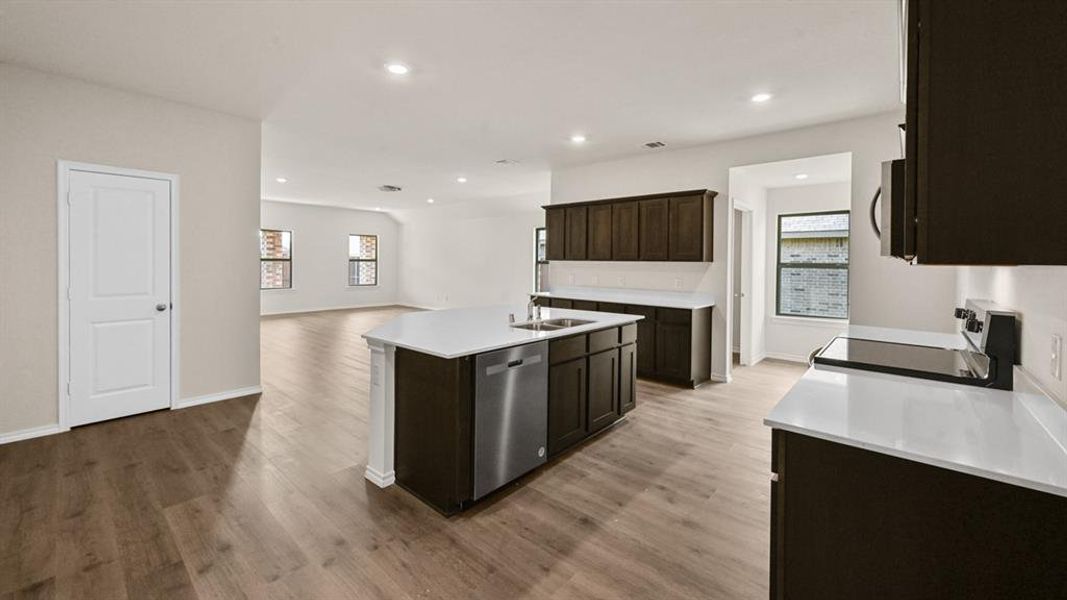 Open-concept kitchen and living area featuring wood-finish flooring, recessed lighting, dark wood-finish cabinetry, light-toned countertops, and a kitchen island with integrated sink and dishwasher Open-concept kitchen and living area featuring wood-finish flooring, recessed lighting, dark wood-finish cabinetry, light-toned countertops, and a kitchen island with integrated sink and dishwasher