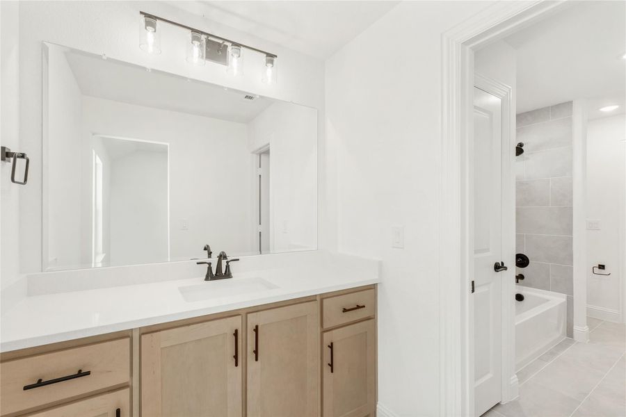 Bathroom with light wood vanity, white countertop, large mirror, and tiled tub-shower combo visible through doorway