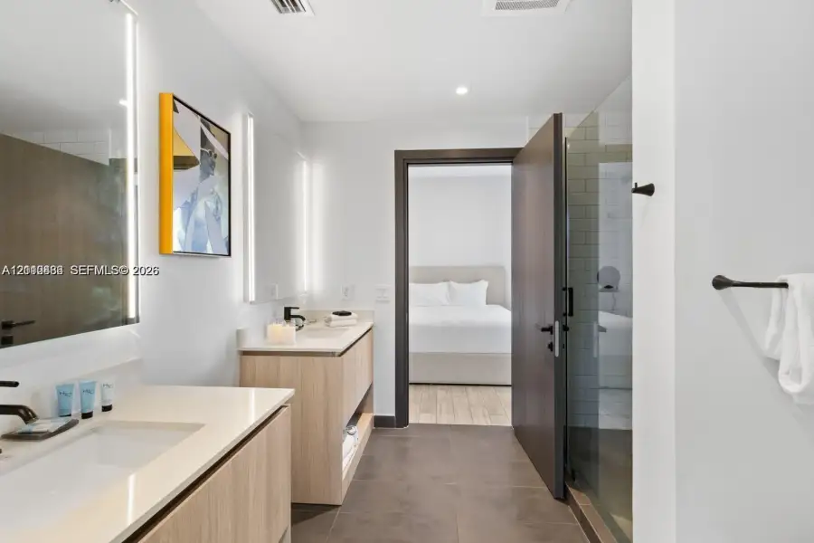 Dual vanity with light wood-finish cabinetry, solid surface countertops, integrated LED-lit mirrors, and glass-enclosed shower with subway tile Dual vanity with light wood-finish cabinetry, solid surface countertops, integrated LED-lit mirrors, and glass-enclosed shower with subway tile