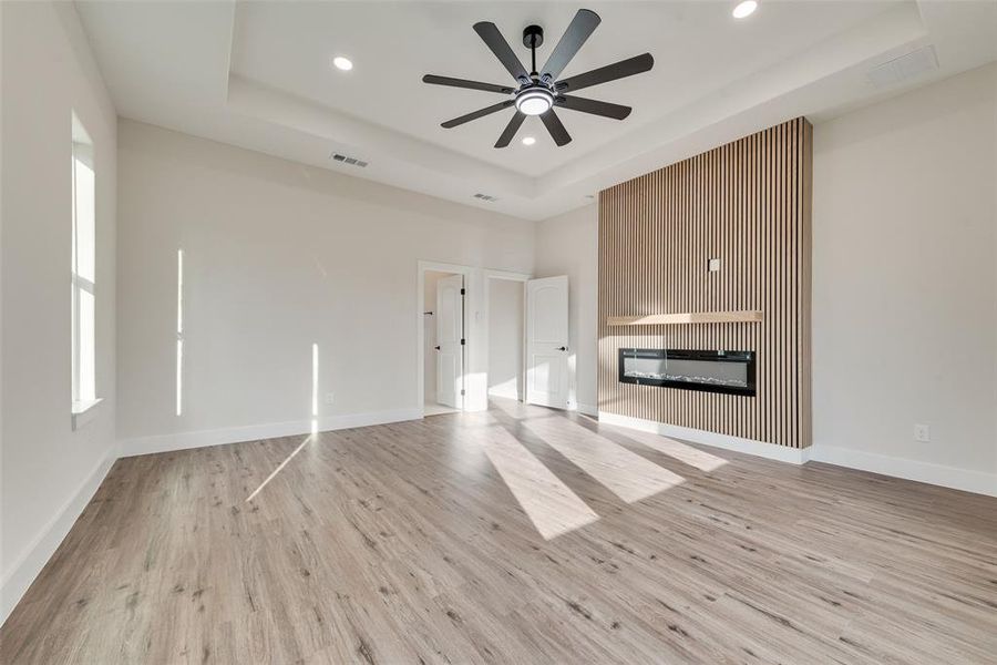 Primary Bedroom featuring a glass covered fireplace, a tray ceiling, light wood finished floors, ceiling fan, and recessed lighting