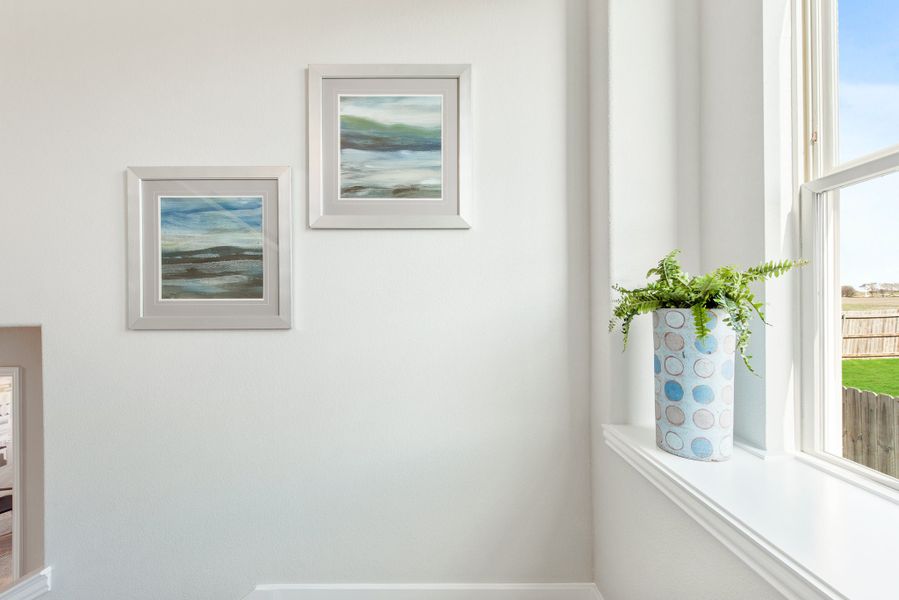 White wall with two framed landscape paintings and a potted fern on a window sill beside a large window
