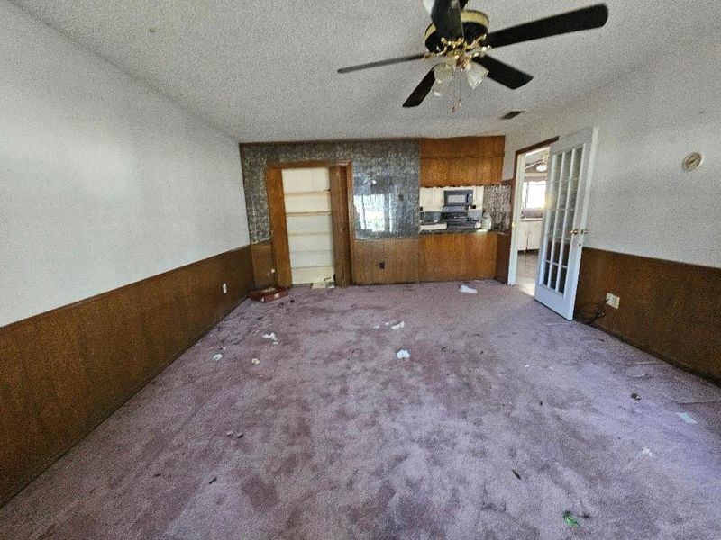 Unfurnished living room with wainscoting, wooden walls, a textured ceiling, dark colored carpet, and a ceiling fan Unfurnished living room with wainscoting, wooden walls, a textured ceiling, dark colored carpet, and a ceiling fan
