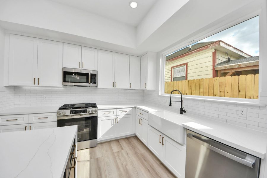 This kitchen features modern white cabinetry, stainless steel appliances, and a spacious farmhouse sink. It has a large window for natural light and sleek countertops, creating a bright and welcoming space.