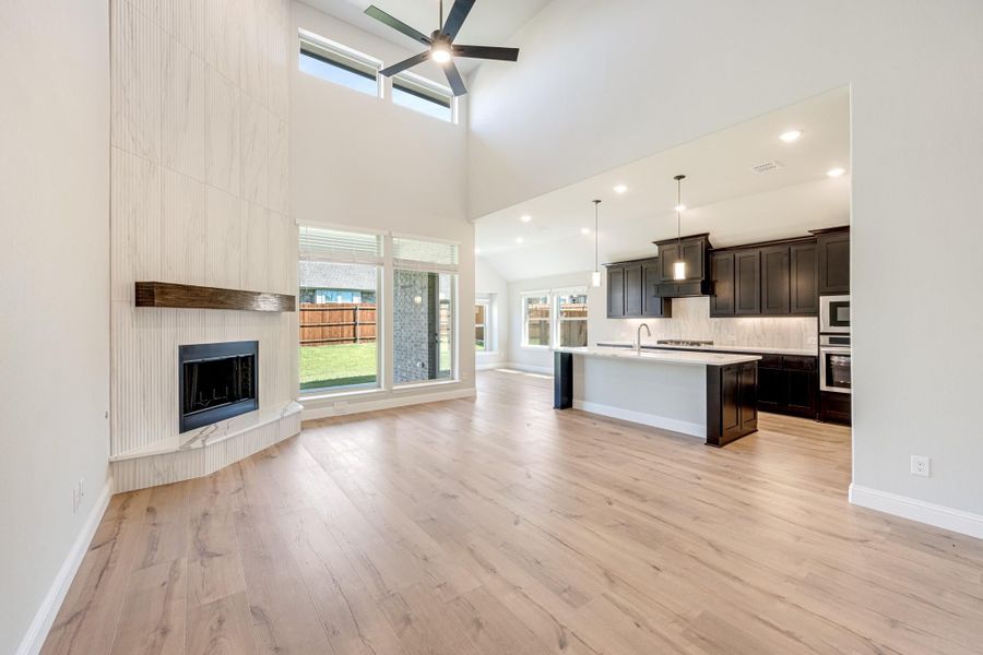 Open living area with tall fireplace, hardwood floors, and open-concept kitchen with dark cabinets and island. Open living area with tall fireplace, hardwood floors, and open-concept kitchen with dark cabinets and island.