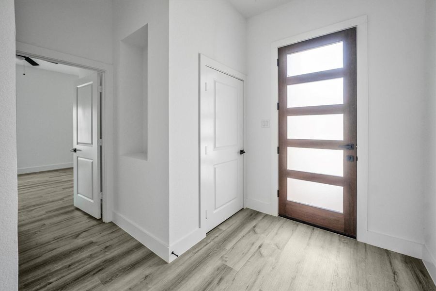 Bright entryway with a modern wooden front door featuring frosted glass panels. It has light flooring, white walls, and a closet for storage. Adjacent room is accessible through a doorway.