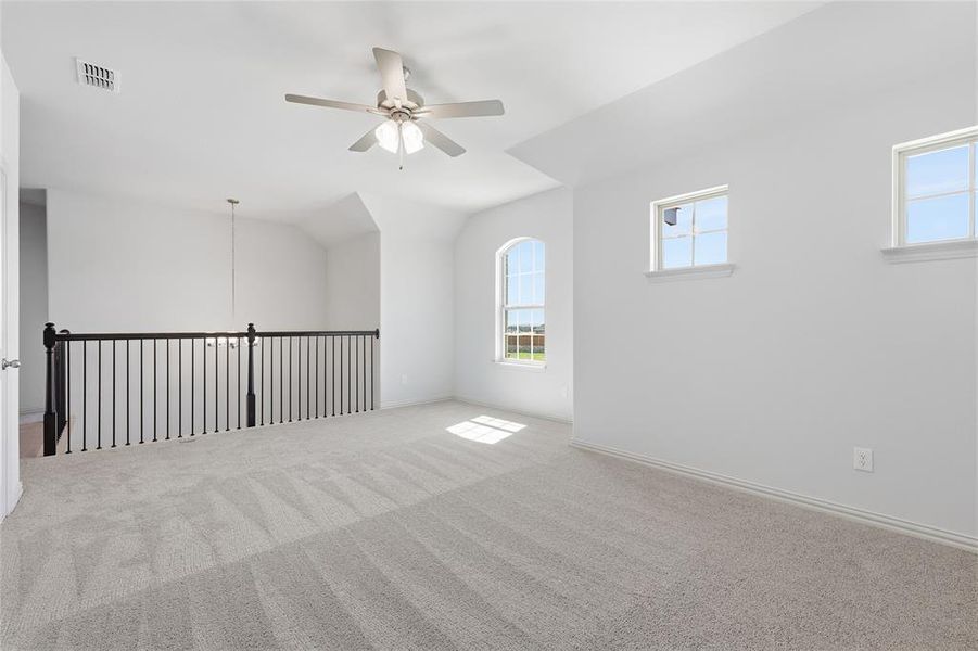 Carpeted open loft space with an arched window, two rectangular windows, a ceiling fan with integrated lighting, and a black wrought iron railing