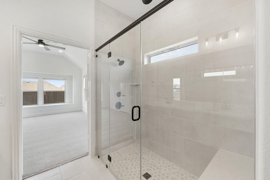 Walk-in glass shower with gray tile walls and black hardware, adjoining carpeted bedroom with ceiling fan visible through doorway Walk-in glass shower with gray tile walls and black hardware, adjoining carpeted bedroom with ceiling fan visible through doorway