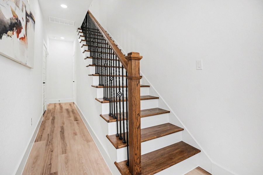 A bright, modern hallway featuring a wooden staircase with elegant iron balusters. The light wood flooring complements the white walls, creating a clean and inviting space.