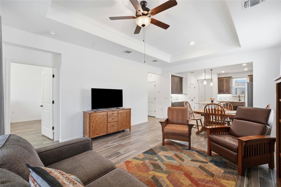 Living area with a raised ceiling, light wood finished floors, ceiling fan, and recessed lighting