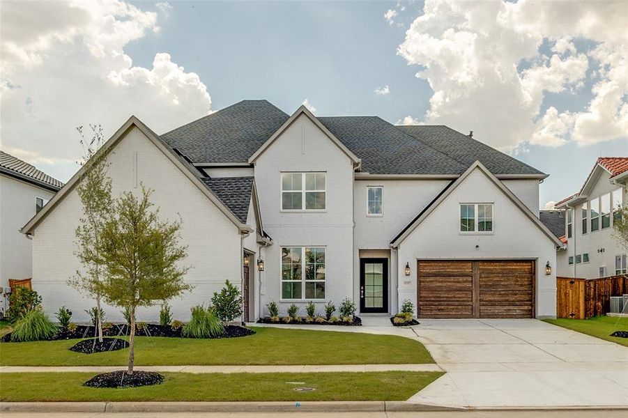 Two-story residence featuring a light-colored brick exterior, dark shingle roof, wood-finish garage doors, and a black front door with glass paneling Two-story residence featuring a light-colored brick exterior, dark shingle roof, wood-finish garage doors, and a black front door with glass paneling