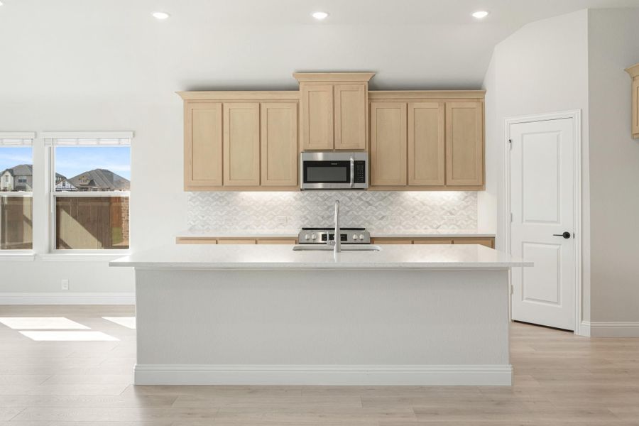 Kitchen with large white island, light wood cabinets, patterned tile backsplash, and stainless steel microwave