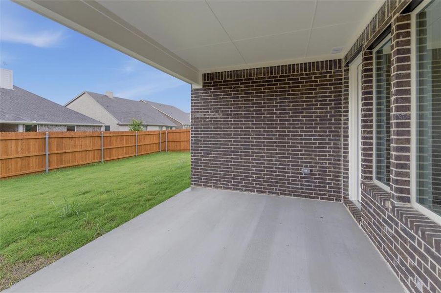 Covered concrete patio featuring a dark brick exterior wall with multiple windows and a private wooden fence enclosing a green lawn