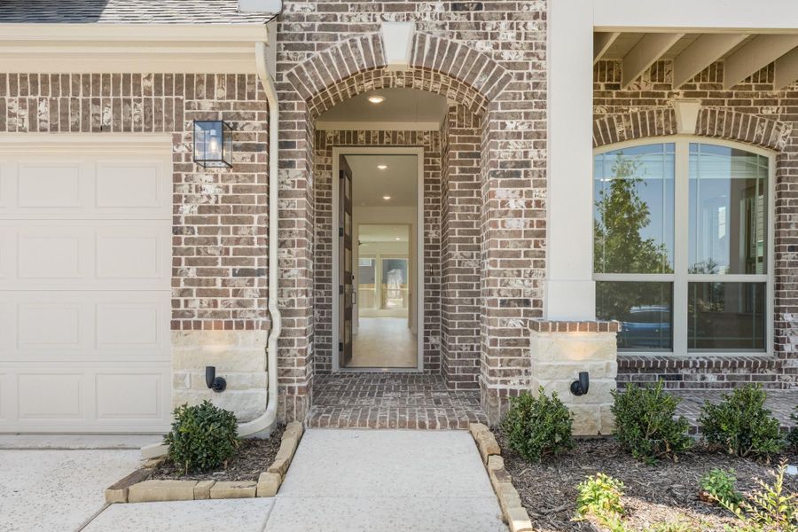 Brick home entrance with arched doorway, stone accents, wall lantern, and white garage door beside front entry