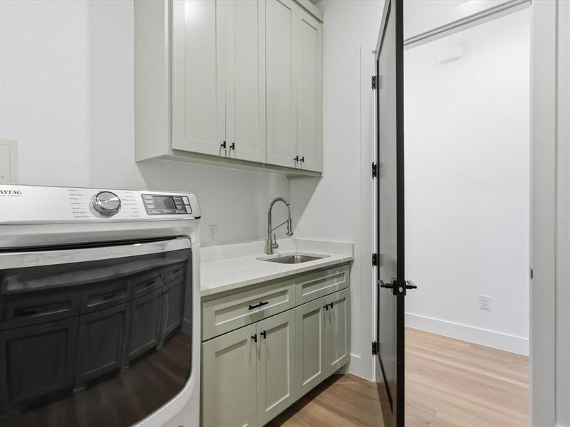 Laundry room featuring light green shaker cabinetry, a stainless steel sink, a light-toned countertop, and wood-finish flooring