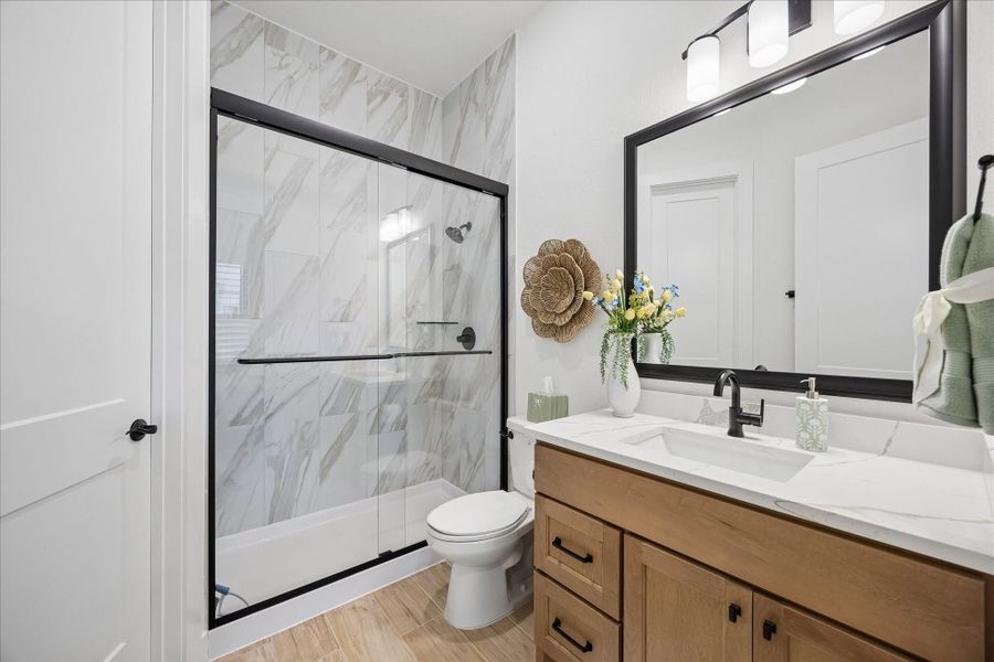 This secondary bathroom has been fully upgraded with wood-plank tile flooring and a spacious walk-in closet. The room features upgraded marble shower tile, oversized granite countertop vanity with soft-close cabinets, an elegant framed mirror, and matte black hardware. The full-size shower is finished with floor-to-ceiling tile and matching matte black fixtures.