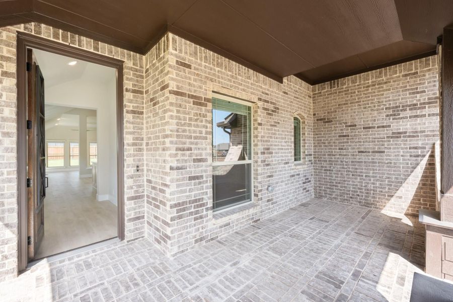 Covered back patio with brick flooring and walls, open door leading to bright interior living area.