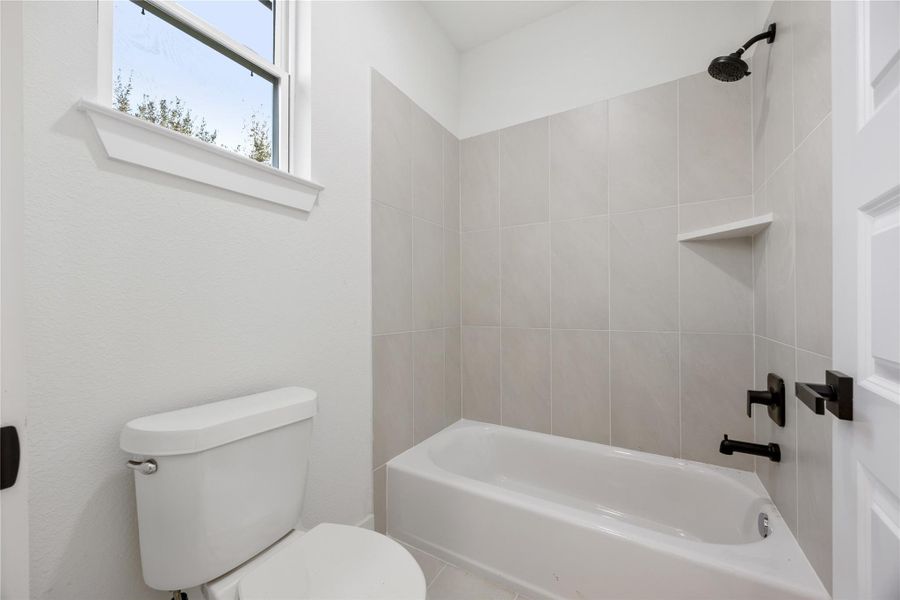 This bathroom features a clean design with neutral tiles, a bathtub-shower combo, black fixtures, and a window providing natural light.