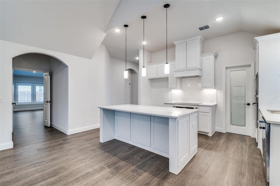 Kitchen with hanging light fixtures, a kitchen island, custom backsplash and pantry door, vaulted ceiling, and white cabinets.