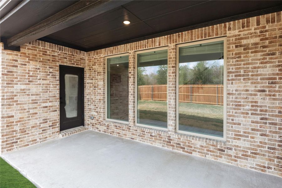 Covered concrete patio with brick exterior wall, large windows, and black door leading into home