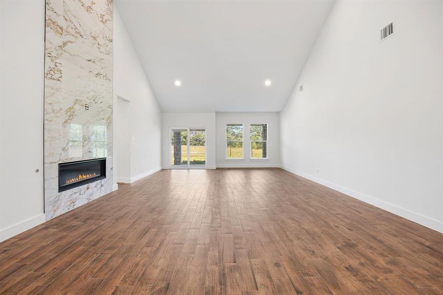Spacious living area featuring a dramatic floor-to-ceiling accent wall with an integrated linear fireplace, wood-finish flooring, soaring vaulted ceilings, and recessed lighting Spacious living area featuring a dramatic floor-to-ceiling accent wall with an integrated linear fireplace, wood-finish flooring, soaring vaulted ceilings, and recessed lighting