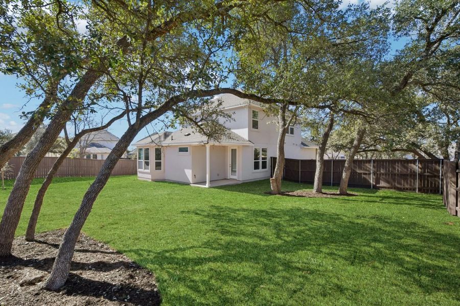 Image of the back exterior of a white two story house with a back covered patio, green grass, a brown wooden fence and multiple trees