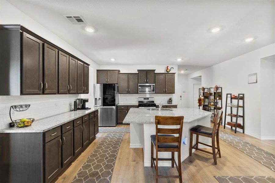 Kitchen featuring dark brown cabinets, light stone counters, stainless steel appliances, a kitchen breakfast bar, and decorative backsplash