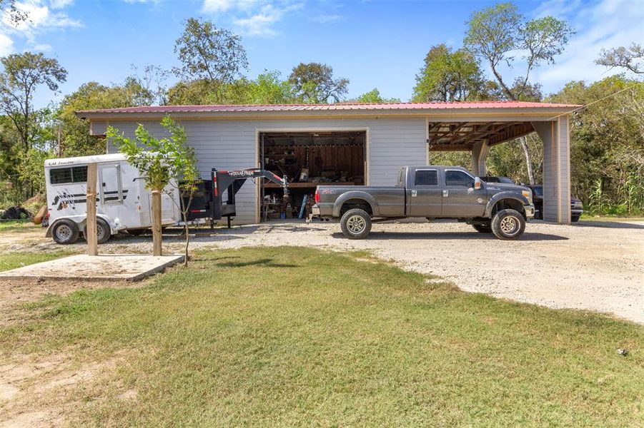 This photo shows a spacious, open-sided barn or workshop with a metal roof, situated on a gravel driveway. It features a large truck parked in front and a horse trailer nearby. The area is surrounded by greenery, indicating a rural setting.