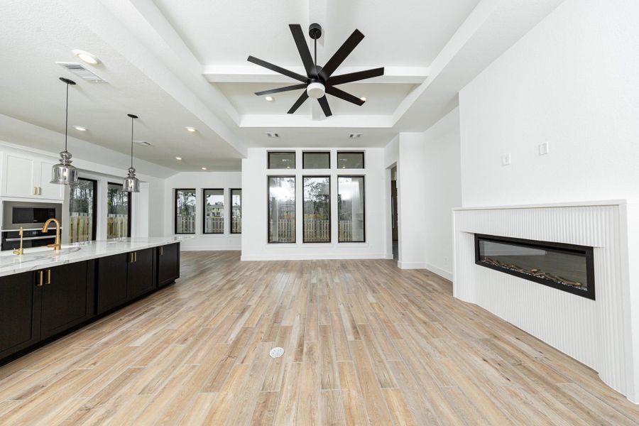 This family room offers a seamless blend of modern luxury and comfortable living within an open-concept design. A sleek electric fireplace is integrated into a modern hearth. An impressive tray ceiling with multiple tiers houses a  contemporary black ceiling fan, providing both comfort and a design statement.