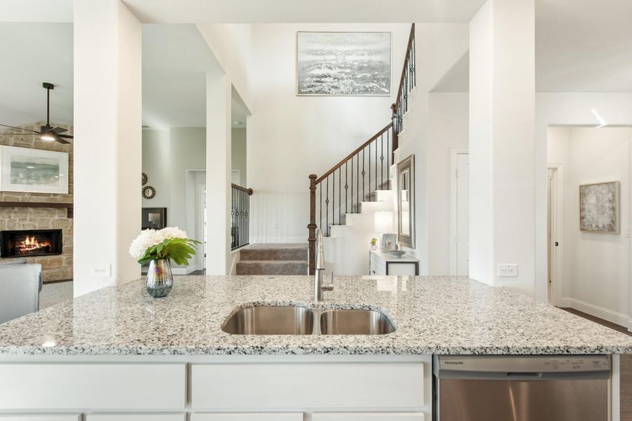 Kitchen island with granite countertop and double sink overlooking open living area with staircase and stone fireplace