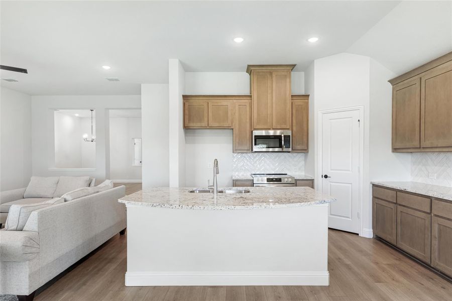 Open kitchen with granite island, tan wood cabinets, white herringbone backsplash, and stainless appliances