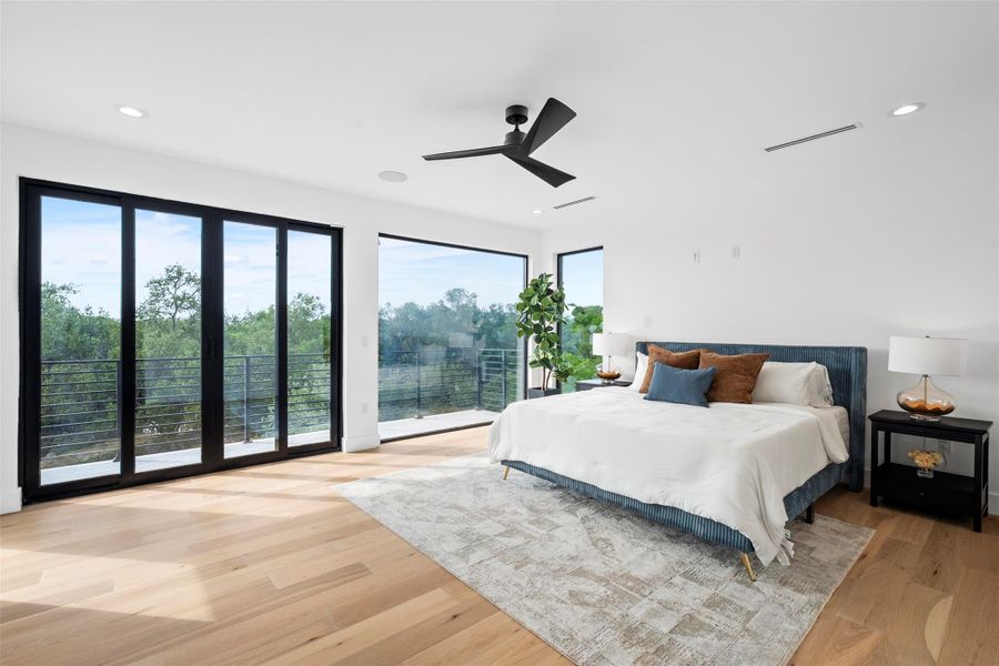 This room features light hardwood floors, a ceiling fan, recessed lighting, and large windows with black frames that offer views of surrounding trees