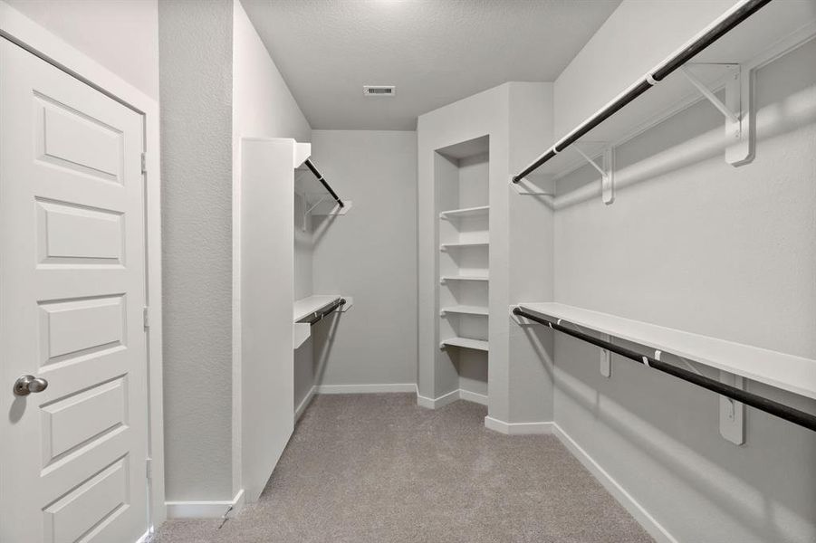 Experience luxury in this spacious walk-in closet with high ceilings and plush carpet. Warm paint tones, built-in shelving, and dark finishes create a contemporary and functional retreat. Sample photo of completed home with similar floor plan. As-built interior colors and selections may vary.