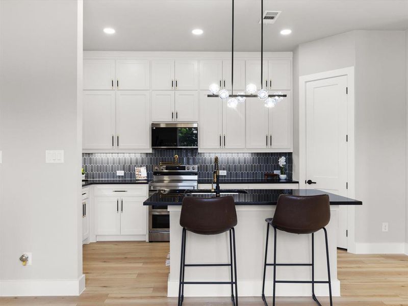 Modern kitchen featuring white shaker cabinetry, stainless steel appliances, a dark tile backsplash, a central island with bar seating, and wood-finish flooring Modern kitchen featuring white shaker cabinetry, stainless steel appliances, a dark tile backsplash, a central island with bar seating, and wood-finish flooring