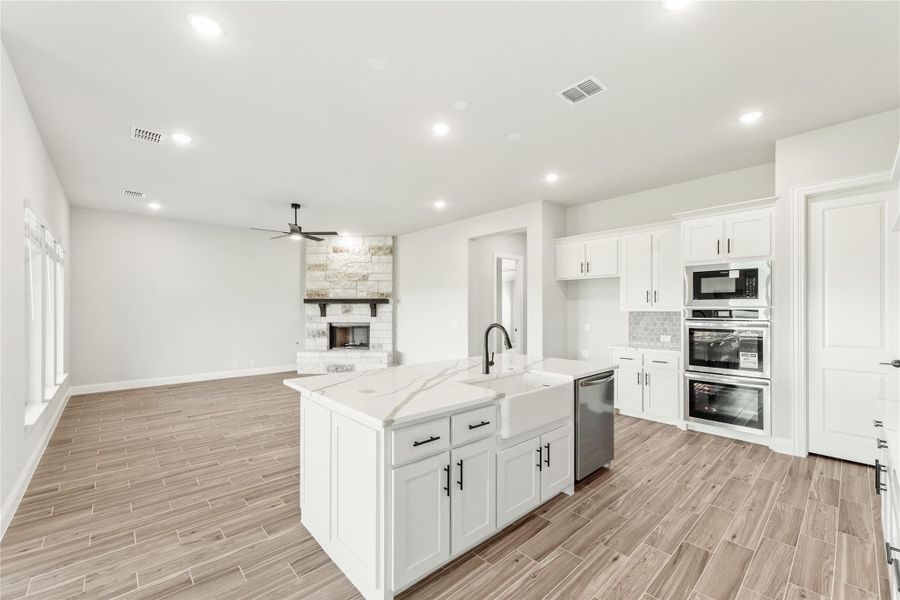 Open-concept kitchen with white cabinets, center island, stainless appliances, and wood-look floors opening to living room with stone fireplace. Open-concept kitchen with white cabinets, center island, stainless appliances, and wood-look floors opening to living room with stone fireplace.