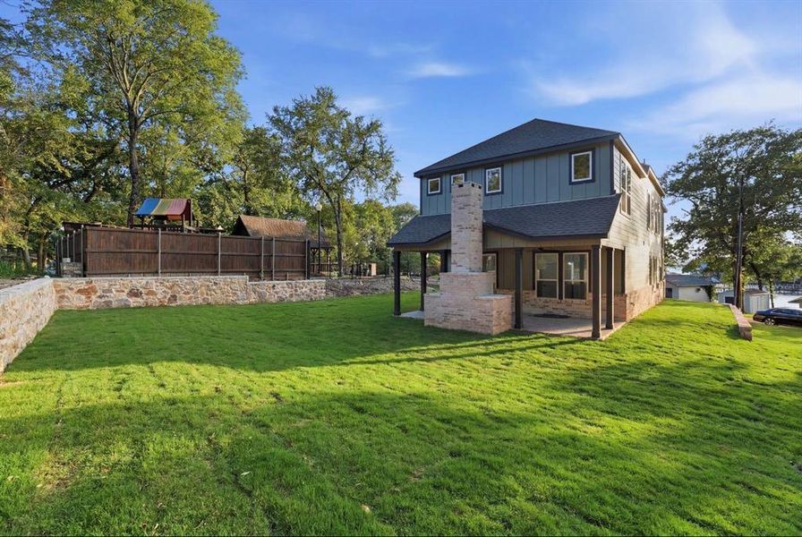 Expansive backyard featuring a stone retaining wall, manicured lawn, and a two-story residence with board and batten siding, multiple windows, and a stone outdoor fireplace with a covered patio Expansive backyard featuring a stone retaining wall, manicured lawn, and a two-story residence with board and batten siding, multiple windows, and a stone outdoor fireplace with a covered patio