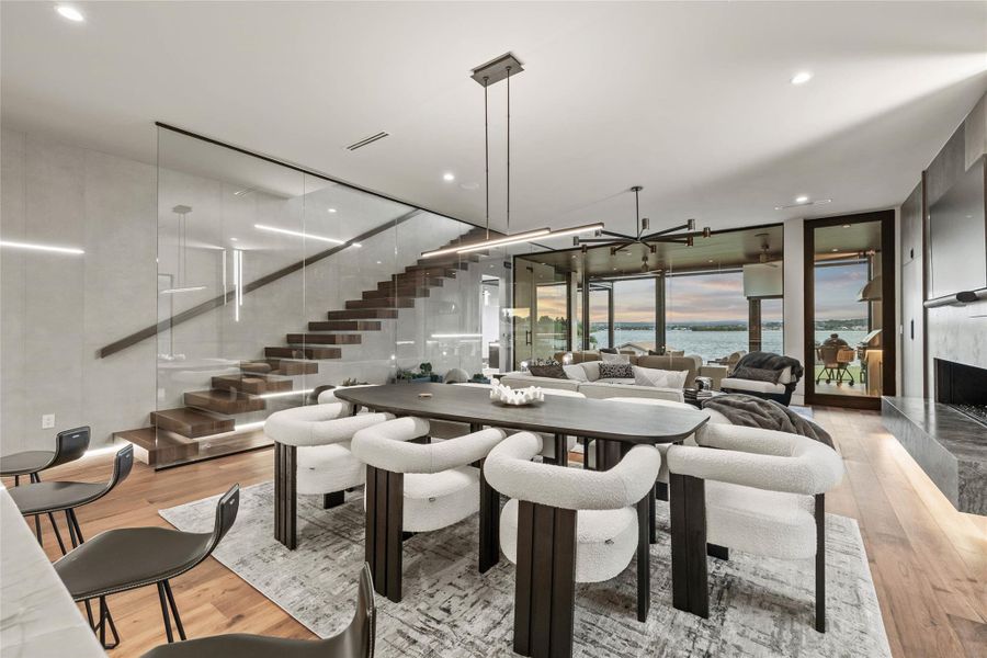 Open-concept living space featuring wood-finish flooring, a glass-paneled staircase with wooden treads, a contemporary fireplace, and floor-to-ceiling windows