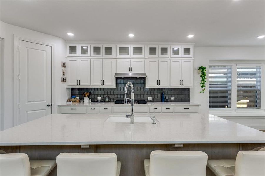 Kitchen featuring glass fronted cabinets, a breakfast bar, light stone counters, a large island with sink, and recessed lighting Kitchen featuring glass fronted cabinets, a breakfast bar, light stone counters, a large island with sink, and recessed lighting