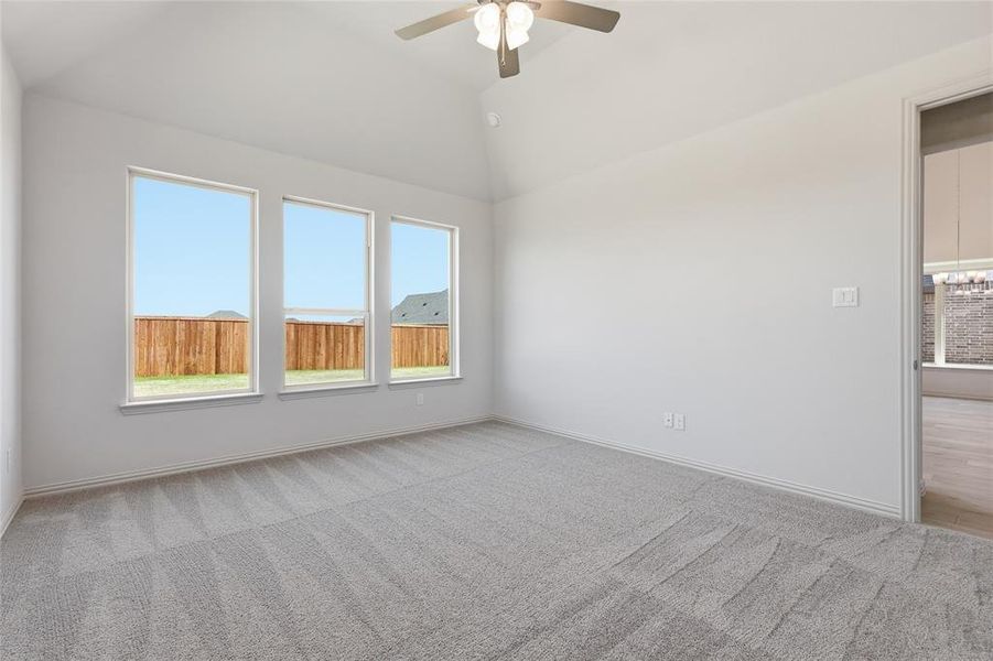 Vaulted ceiling room featuring three expansive windows, light gray patterned carpeting, a ceiling fan with integrated lighting, and a white baseboard trim