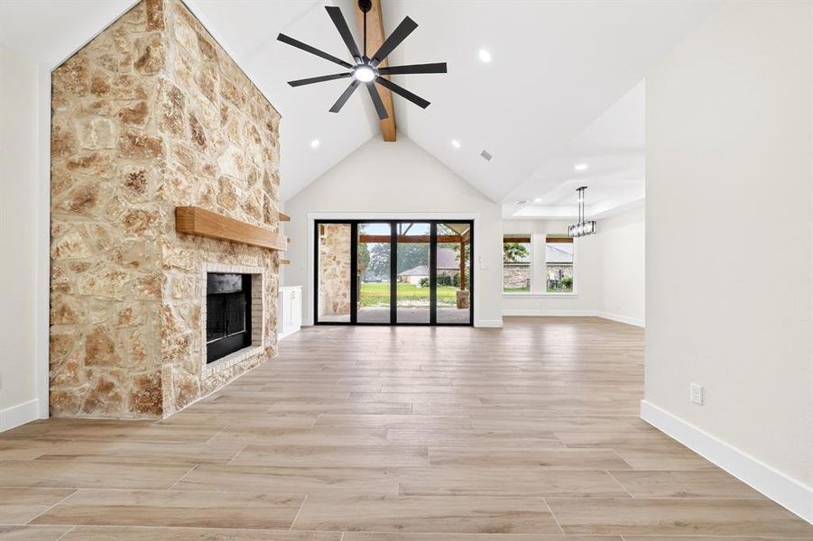 Vaulted ceiling great room featuring a full-height stone fireplace with a wood mantle, wood-finish flooring, a ceiling fan, and large sliding glass doors opening to an exterior covered patio Vaulted ceiling great room featuring a full-height stone fireplace with a wood mantle, wood-finish flooring, a ceiling fan, and large sliding glass doors opening to an exterior covered patio