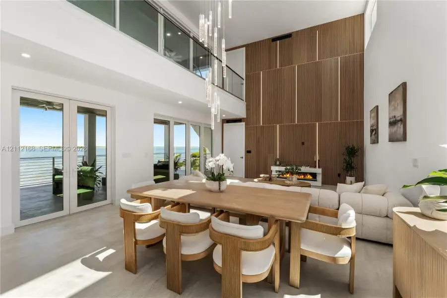 Double-height living area featuring floor-to-ceiling wood paneled accent wall with integrated linear lighting, glass-railed mezzanine, contemporary multi-pendant light fixture, and multiple sets of glass panel doors Double-height living area featuring floor-to-ceiling wood paneled accent wall with integrated linear lighting, glass-railed mezzanine, contemporary multi-pendant light fixture, and multiple sets of glass panel doors
