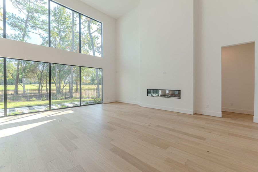 Bright, spacious living room with floor-to-ceiling windows offering scenic views and abundant natural light. Features light wood flooring and a modern, built-in linear fireplace. High ceilings add to the open feel.