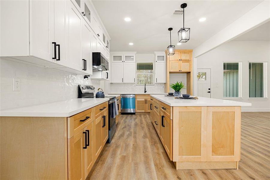 This modern kitchen features sleek white cabinetry paired with natural wood accents, a spacious island with ample seating, stainless steel appliances, and stylish pendant lighting. The open layout and large windows ensure plenty of natural light, enhancing the inviting atmosphere.