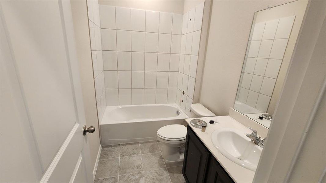 Full bathroom featuring a white tub and shower surround with white tiled walls, a white toilet, and gray tiled flooring Full bathroom featuring a white tub and shower surround with white tiled walls, a white toilet, and gray tiled flooring