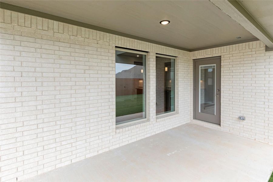 Covered back patio with concrete floor, white brick exterior, large windows, and gray back door with glass panel
