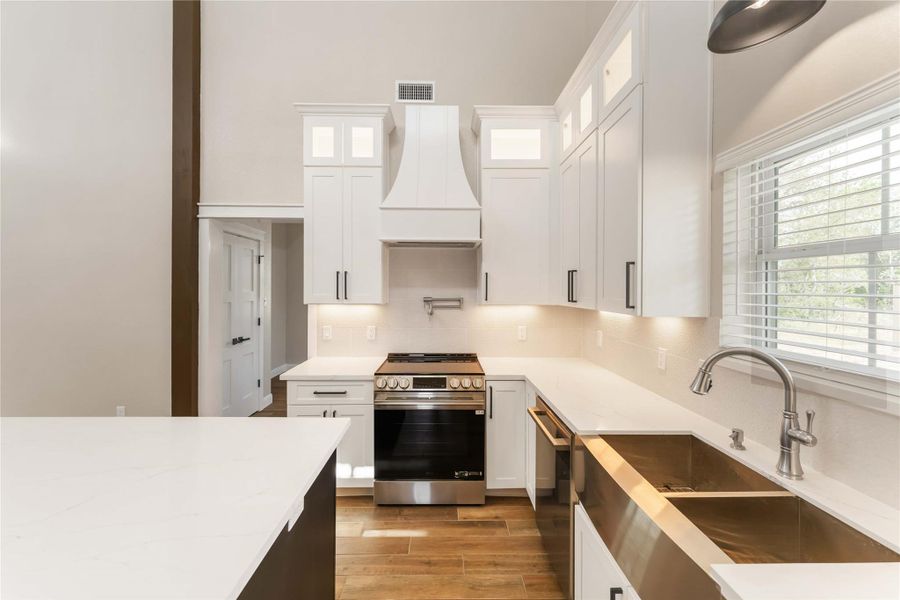 This kitchen features a modern design with high ceilings, white cabinetry, and sleek countertops. It includes stainless steel appliances, a deep sink, and ample natural light from a large window.