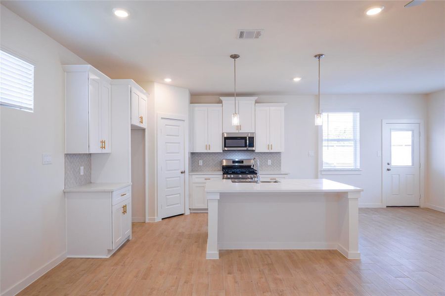 Bright, modern kitchen with white cabinetry, stainless steel appliances, and a large island. Features pendant lighting, ample counter space, and natural light from multiple windows.
