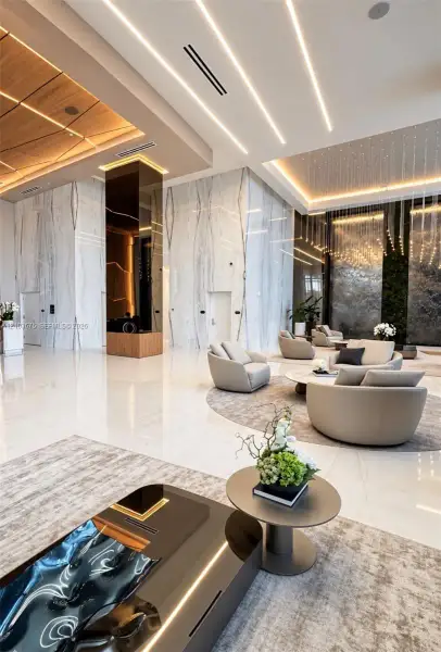 High-ceilinged lobby featuring large-format polished tile flooring, full-height stone wall cladding, integrated linear recessed lighting, and a tiered wood-panel ceiling feature High-ceilinged lobby featuring large-format polished tile flooring, full-height stone wall cladding, integrated linear recessed lighting, and a tiered wood-panel ceiling feature